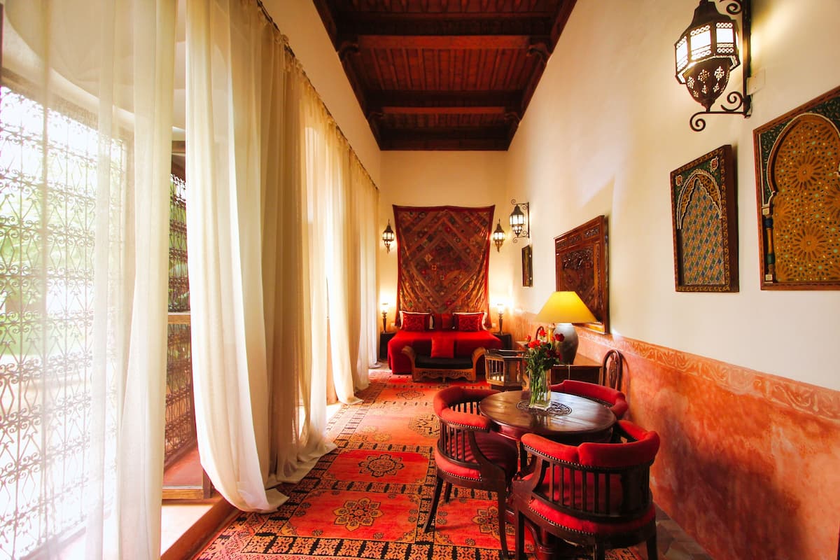 A room with a patterned carpet, a sofa, a dining table with chairs, and wall-mounted lamps.