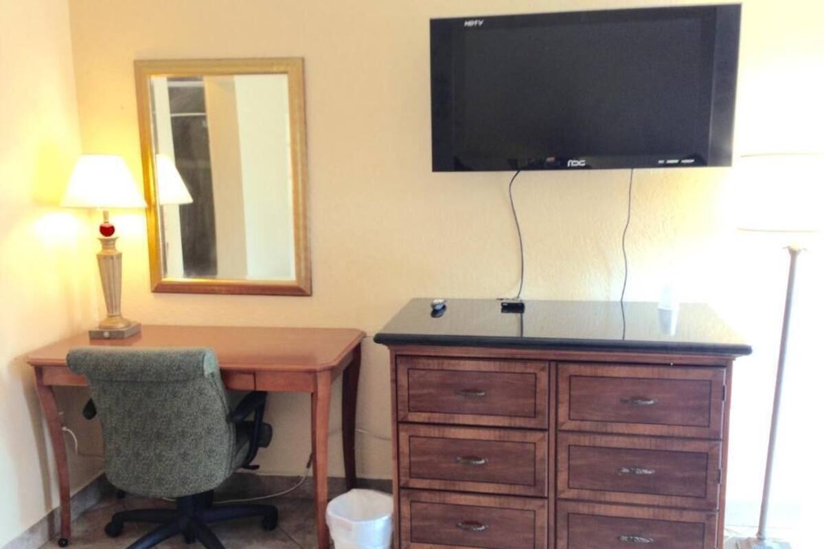 A hotel room with a wooden desk, a chair, a television mounted on the wall, a mirror, and a trash bin.