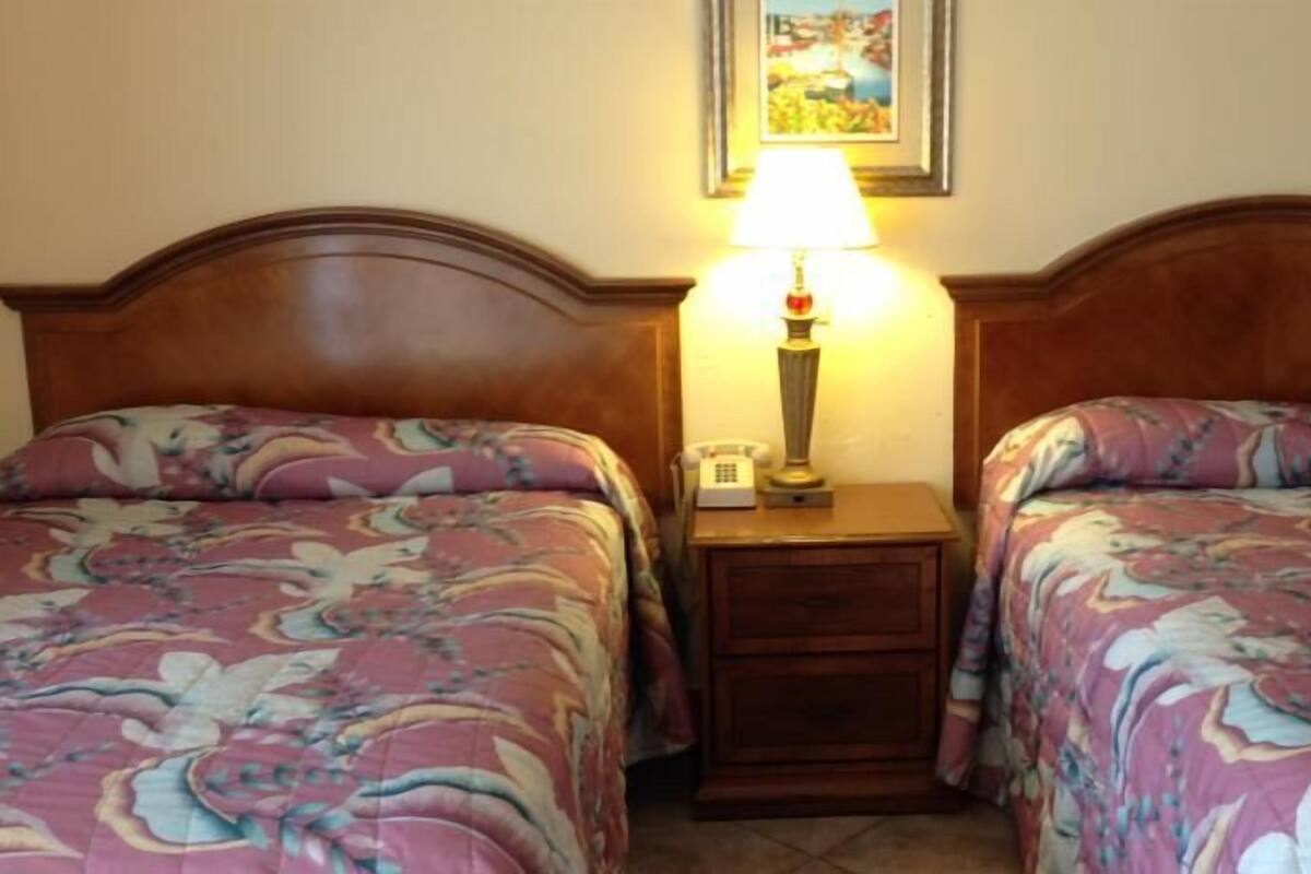 A hotel room with two beds, each with floral-patterned bedding. There is a nightstand between the beds with a lamp and a telephone.