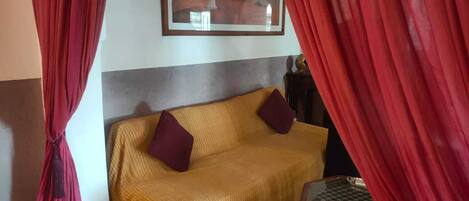 A room with a yellow sofa, red curtains, a framed picture on the wall, and a small wooden table.