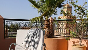A balcony with a lounge chair, a small table with breakfast items, a potted plant, and a view of a building with a tower. - Riad Marelia (Marrakech)