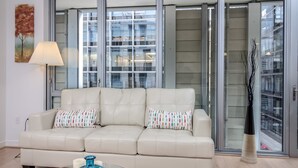 A modern living room with a beige leather sofa, two patterned pillows, a glass coffee table with decorative items, a floor lamp, and a large window with a view of a cityscape. - Arlington Fully Furnished Apartments (Arlington)