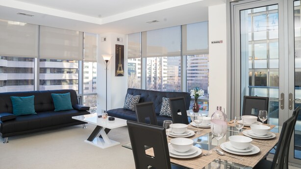 A modern living room with a black sofa, a glass dining table set for four, and large windows offering city views. - Arlington Fully Furnished Apartments (Arlington)
