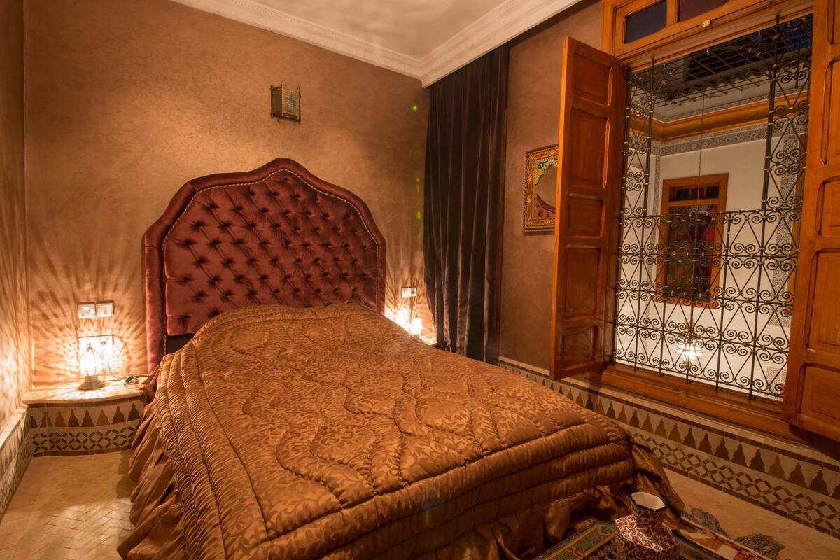 Un dormitorio con una cama grande, cabecera de madera, una ventana con rejas decorativas y una mesita de noche con lámpara.