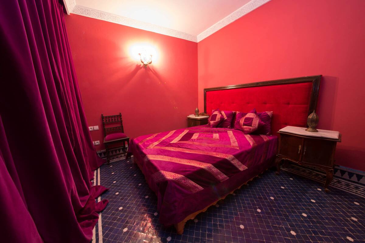 Una habitación con pared roja, una cama con cabecera roja, una mesita de noche y una lámpara fijada a la pared.