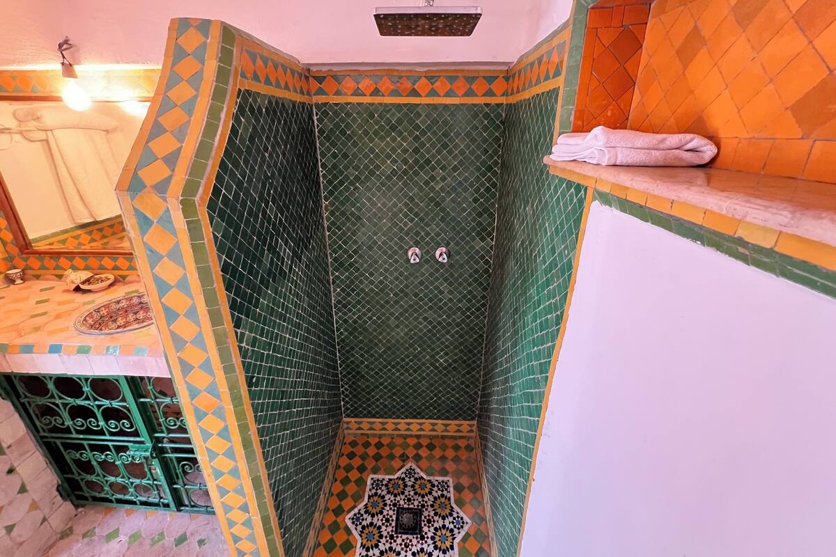 Un baño con zona de ducha con azulejos estampados en tonos verde y naranja, un lavabo con un espejo decorativo y un perchero.