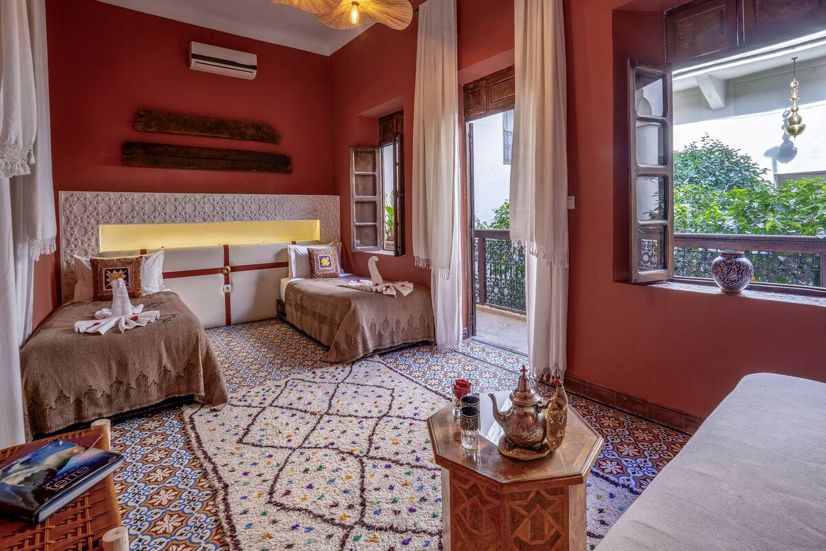 Una habitación con dos camas, una lámpara decorativa en el techo, un ventanal con vistas a la vegetación y una pequeña mesa con un florero.