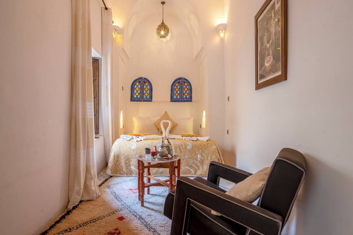 Un dormitorio con cama, una silla, una mesa con tetera y dos ventanas decorativas.