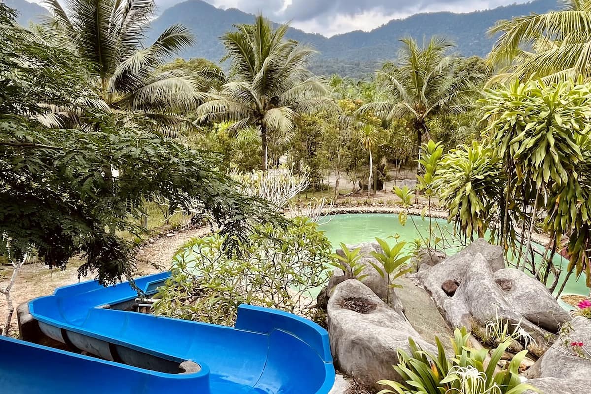 A blue slide in a tropical garden with a pool and mountains in the background.