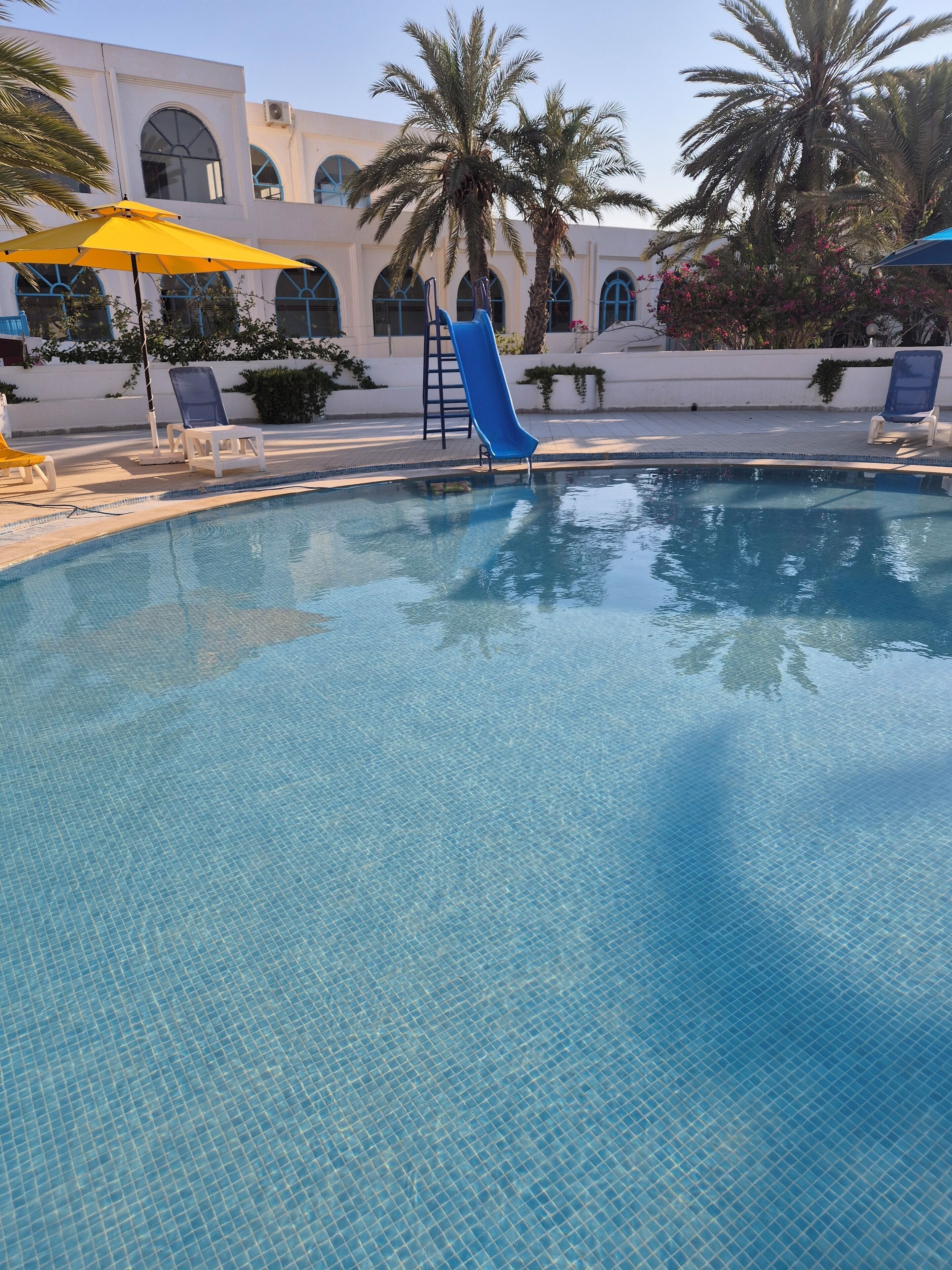 A swimming pool with a blue slide, lounge chairs, and palm trees.
