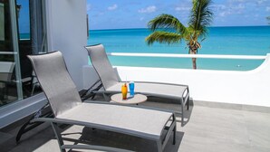 Een balkon met twee loungestoelen, een klein rond tafeltje met drankjes en uitzicht op de oceaan en palmbomen.