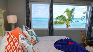 Een slaapkamer met een bed, kussens, een lamp en uitzicht op de oceaan door een raam.