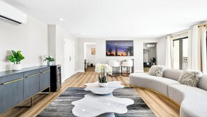 Una sala de estar moderna con un sofá gris, una mesa de centro blanca y un mueble gris. Hay un cuadro en la pared y una planta sobre el mueble.
