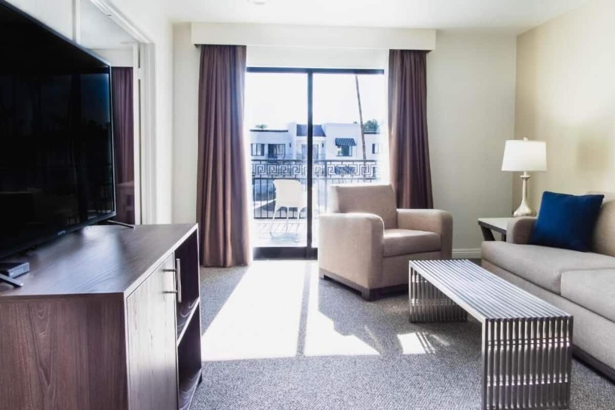 Una habitación de hotel moderna con televisión de pantalla plana, un sofá, una silla, una mesita y un balcón con cortinas.