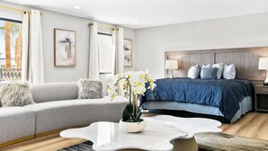 Una habitación de hotel moderna con un sofá gris, una cama con edredón azul, una mesa de centro blanca y un ventanal con cortinas.