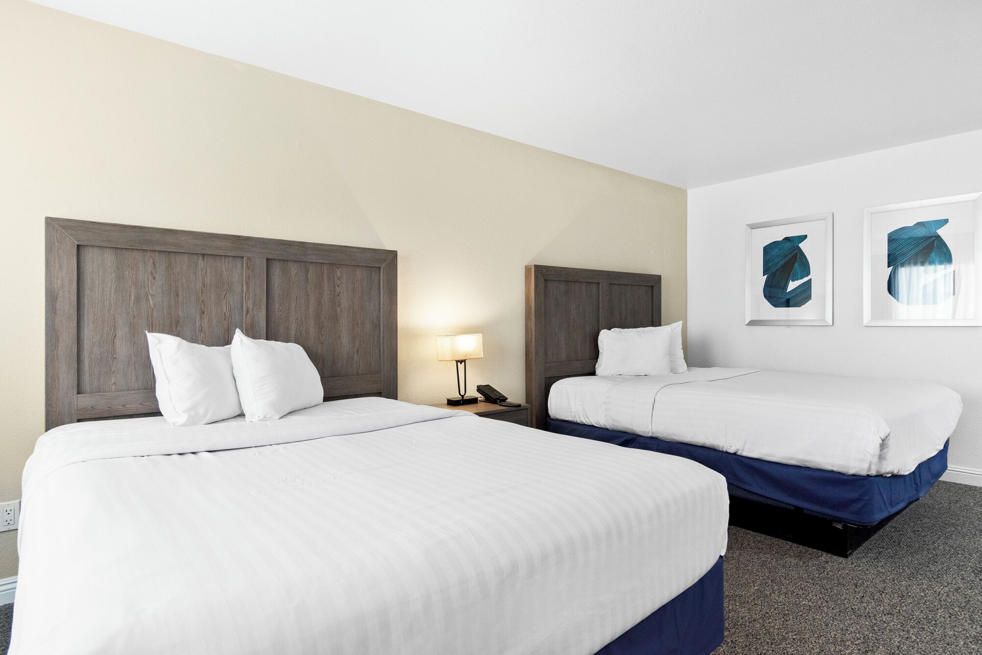 A hotel room with two beds, a nightstand with a lamp, and two framed artworks on the wall.