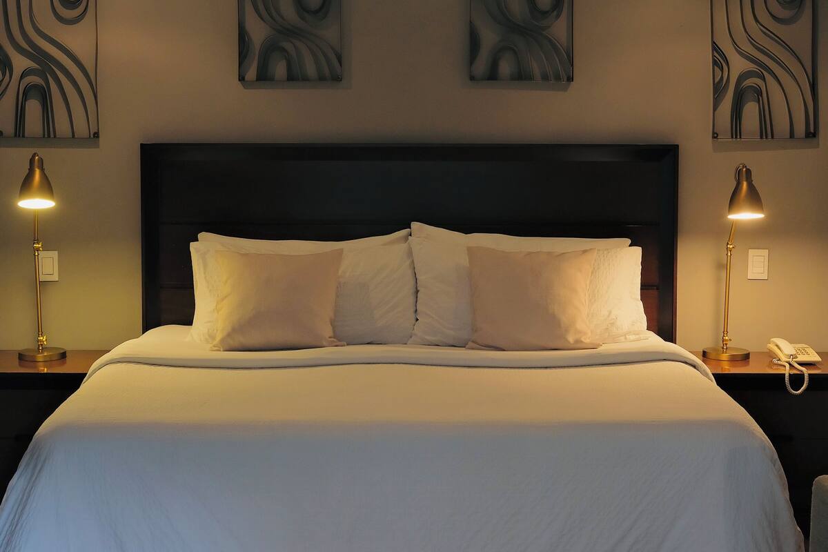 Una cama bien hecha con sábanas blancas, dos lámparas de noche y una obra de arte abstracto en la pared.