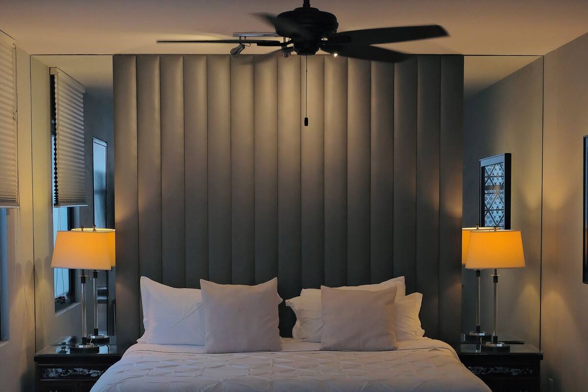 Un dormitorio moderno con una cama grande, dos mesitas de noche con lámparas, un ventilador de techo y un espejo grande en la pared.