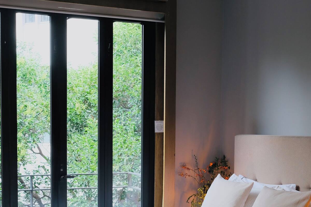 Un dormitorio con una cama grande, una ventana con vista a la vegetación y una mesita de noche con un florero.