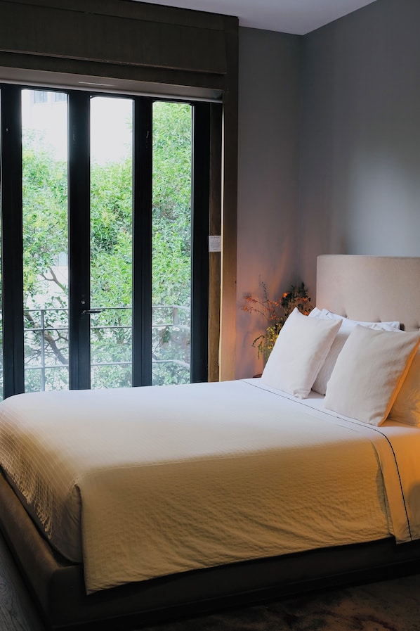 A bedroom with a large bed, a window with a view of greenery, and a bedside table with a vase.