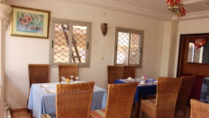 Un comedor con sillas y mesas de mimbre, un cuadro en la pared y ventanas con contraventanas decorativas.