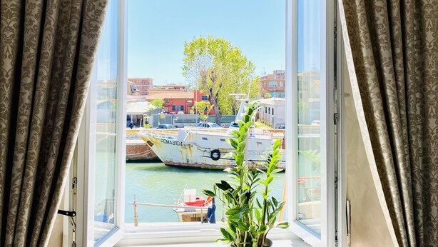 A view from a window with curtains, featuring a marina with boats and buildings in the background. - Domus Valadier Guesthouse (Fiumicino)