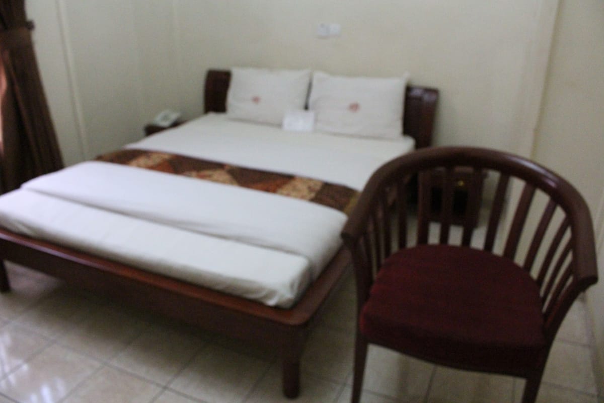 Uma cama com estrutura de madeira, colchão branco e manta com padrão marrom e branco. Há uma cadeira de madeira com almofada escura ao lado da cama.