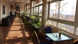 Un pasillo de hotel con una fila de plantas en macetas, una mesa con un florero y vistas al exterior a través de grandes ventanales.