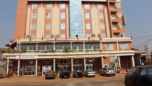 Un edificio de varios pisos con un eje central para ascensor de vidrio, locales comerciales con los rótulos "AfriLand First Bank", “Bar-Restaurant” y “Hôtelage”. Varios automóviles están estacionados en la parte delantera.