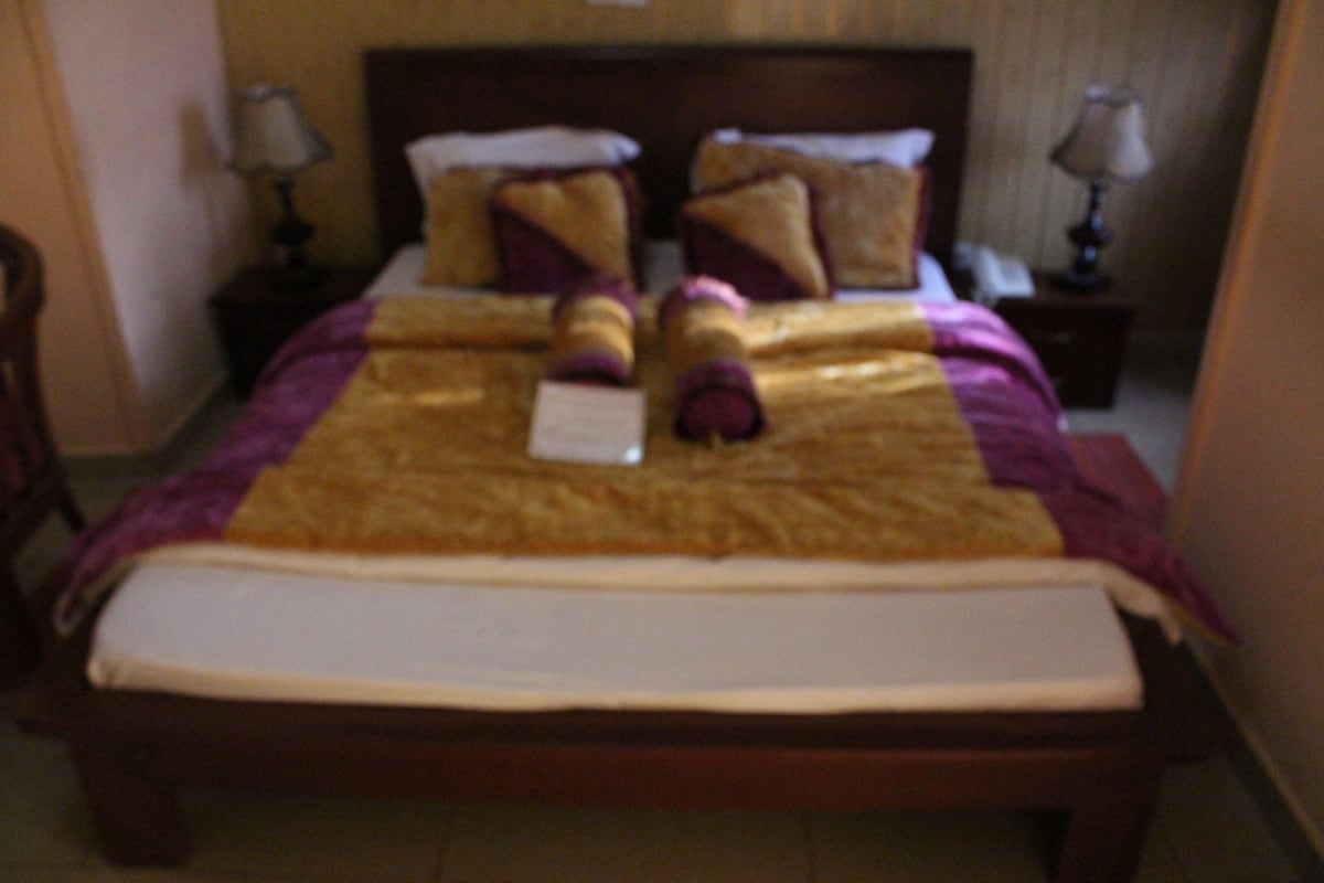 Quarto de hotel com cama, almofadas, mesa de cabeceira e abajur.
