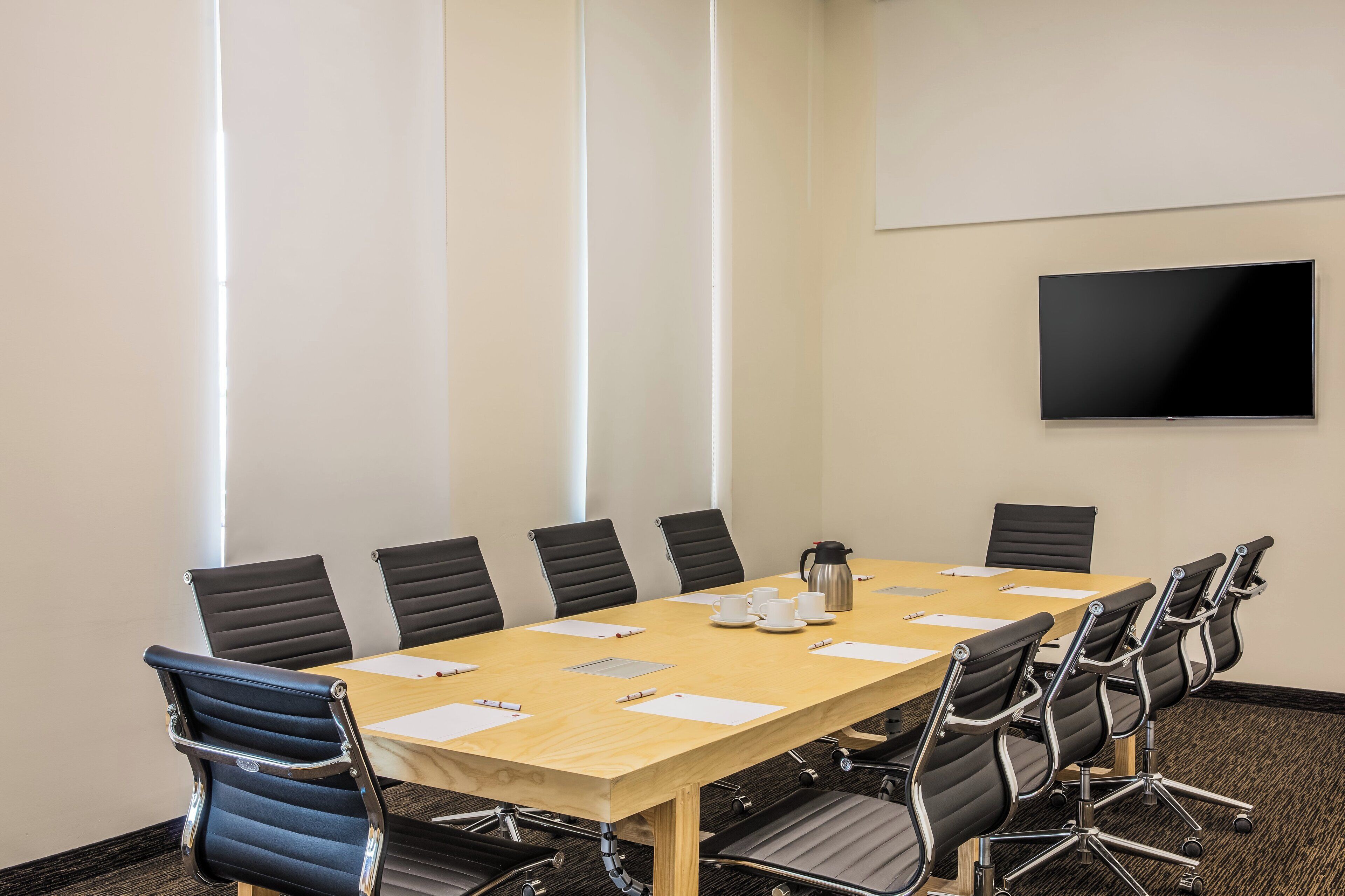 A conference room with a long table, black chairs, a coffee pot, and a flat-screen TV on the wall.