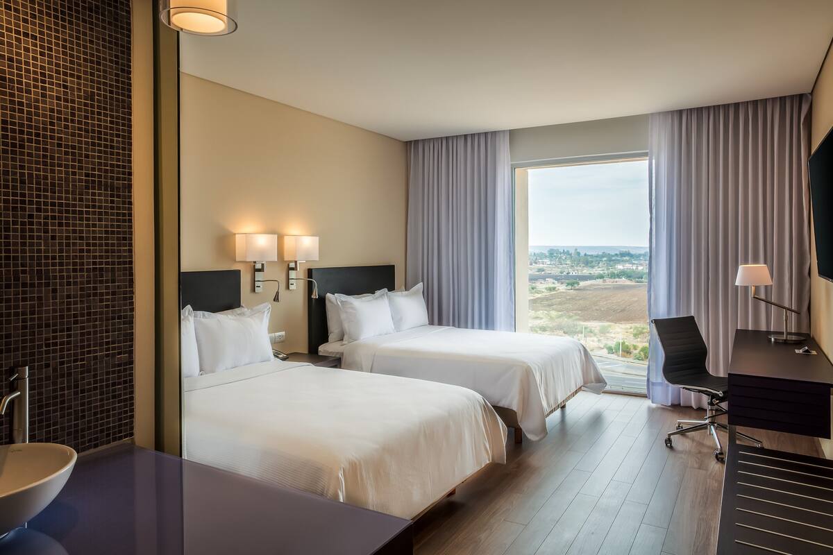 A hotel room with two beds, a desk, a chair, a large window with a view, and a tiled wall.