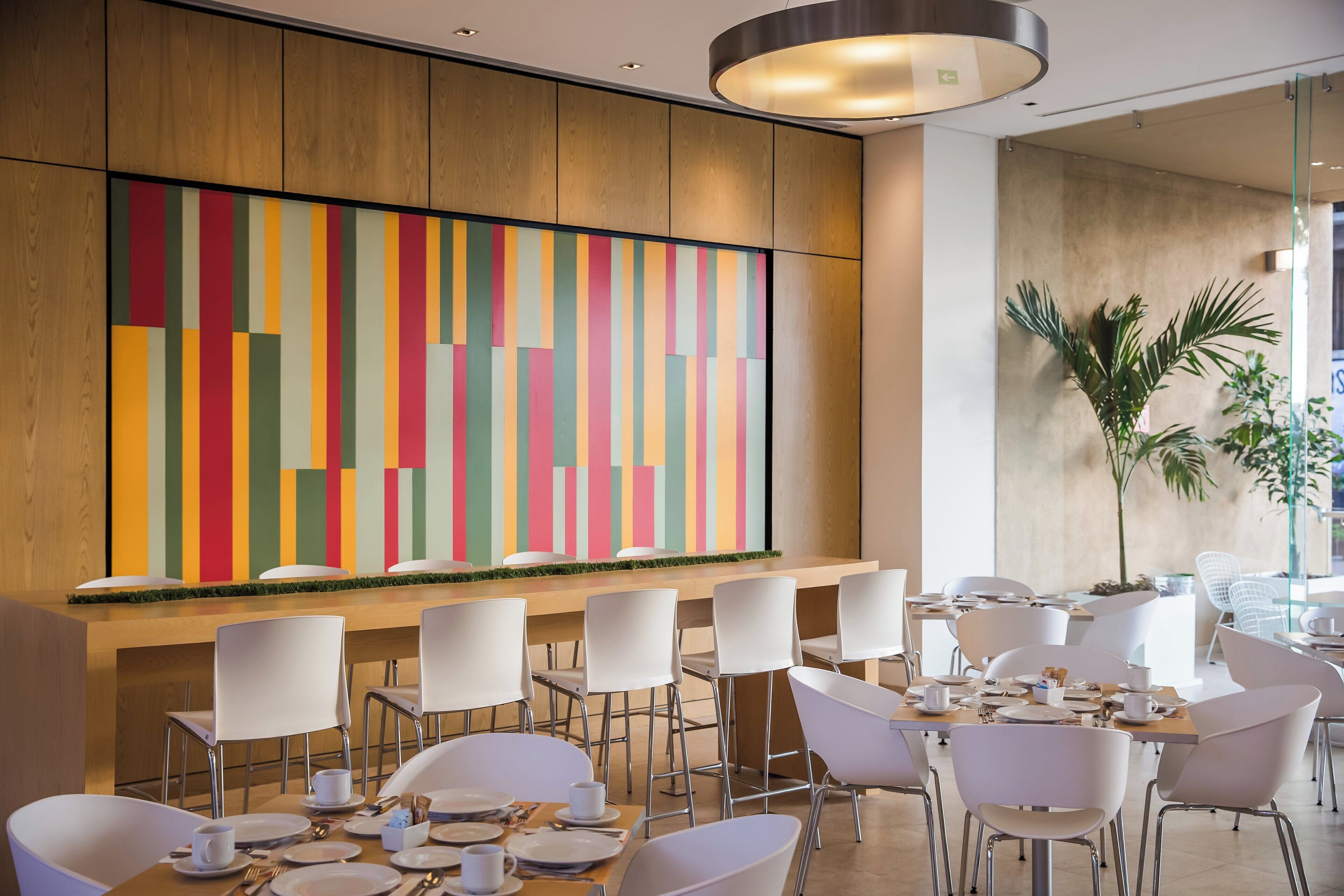 A modern dining area with white chairs and tables, a colorful abstract wall, and a potted plant.