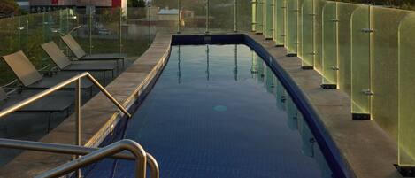 A swimming pool with glass railings, a metal ladder, and lounge chairs.