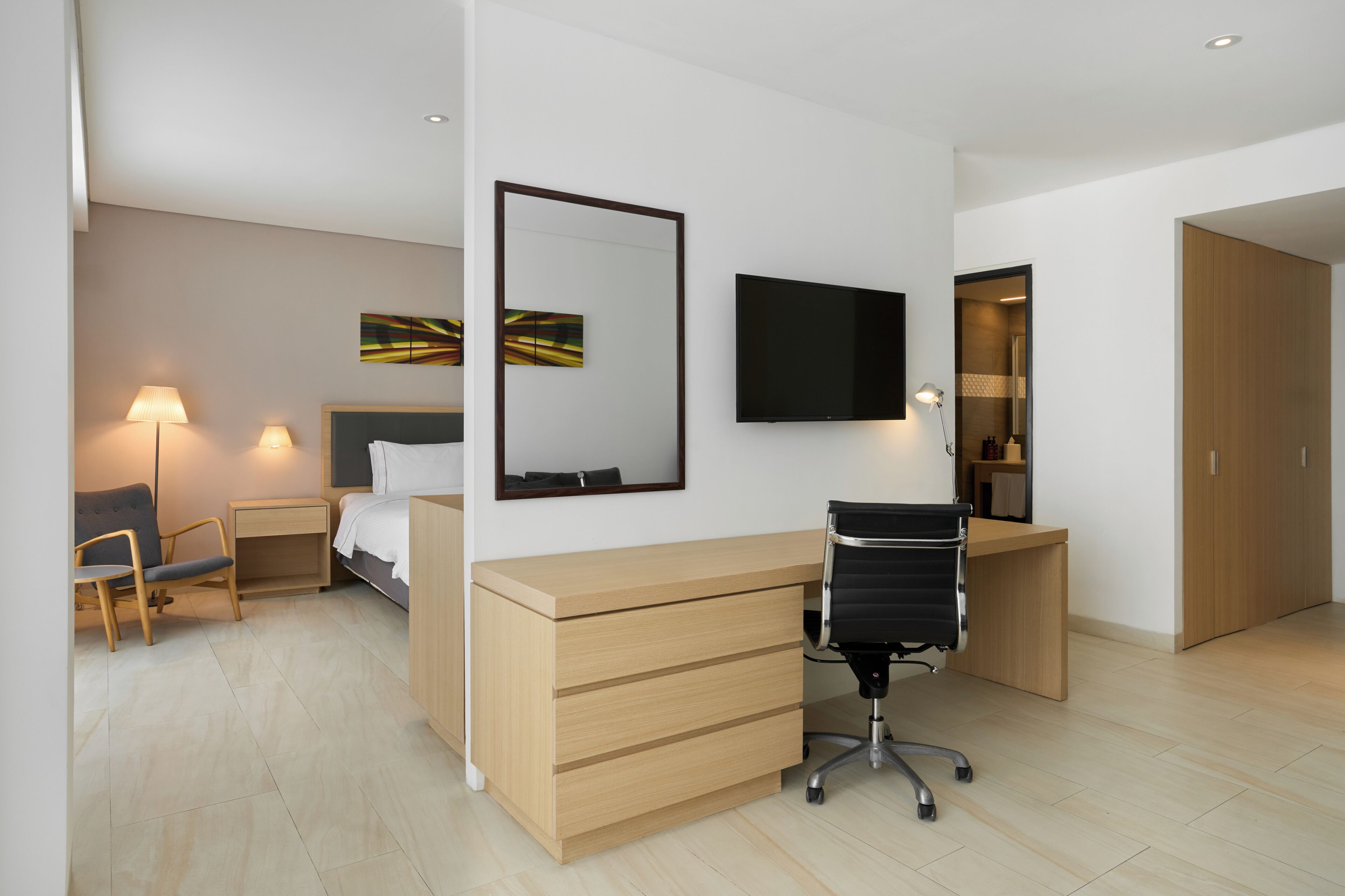 A modern hotel room with a bed, desk, chair, TV, and artwork on the wall.