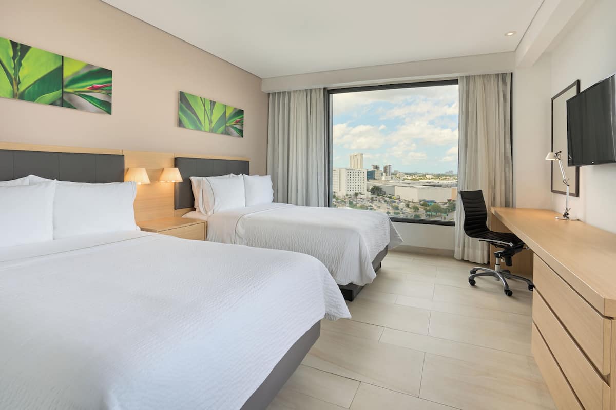Habitación de hotel con dos camas, un escritorio, una silla, un televisor y un ventanal con vista a la ciudad.