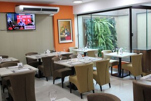 A restaurant with tables set for dining, a television displaying a news program, and a large window with a view of a tropical plant. - Howard Johnson by Wyndham Ciudad del Este (Ciudad Del Este)