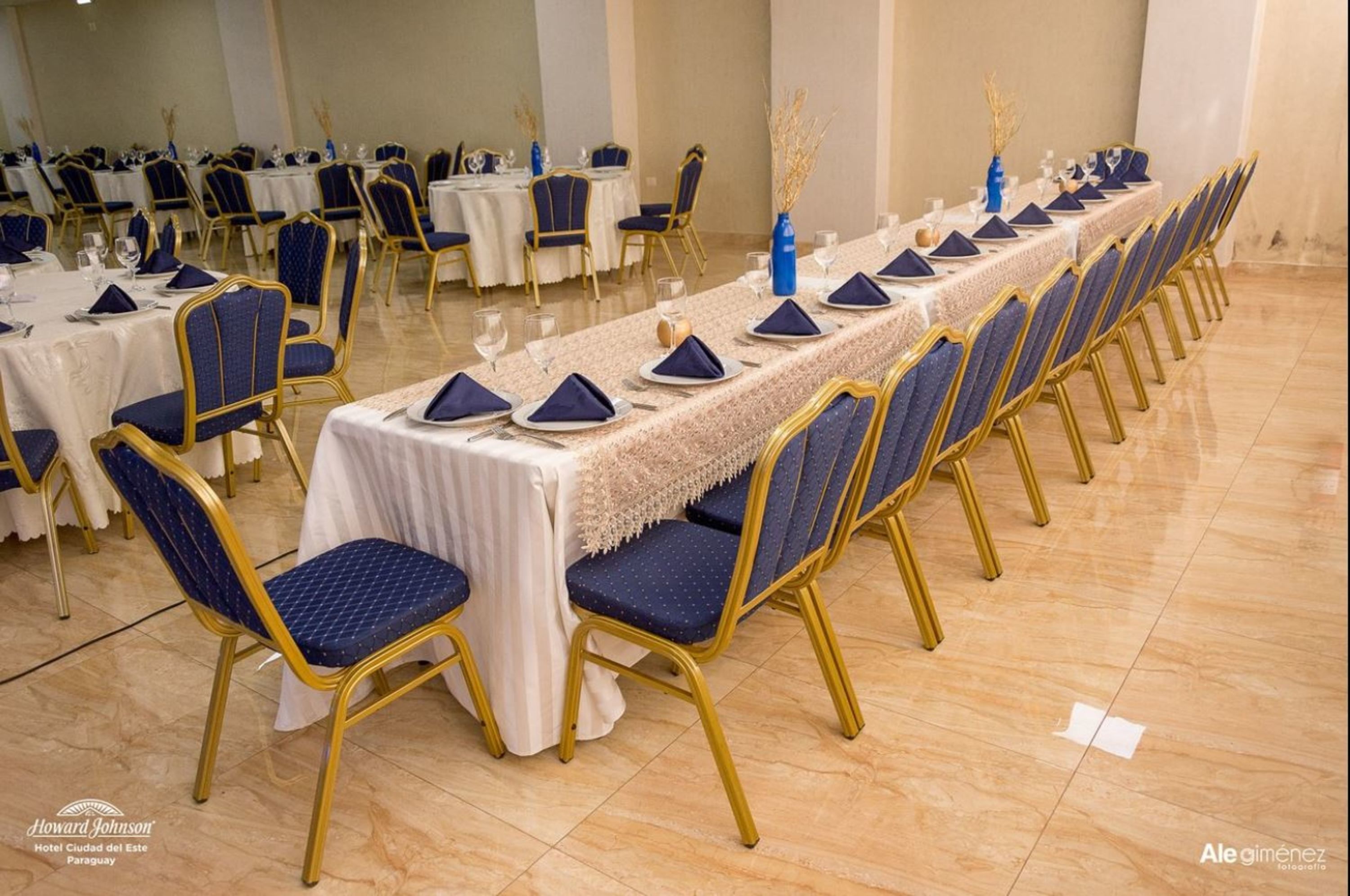 A banquet hall with long tables set for a formal event, featuring gold chairs and white tablecloths.