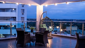 A rooftop terrace with wicker furniture, a hanging chair, and a city view.