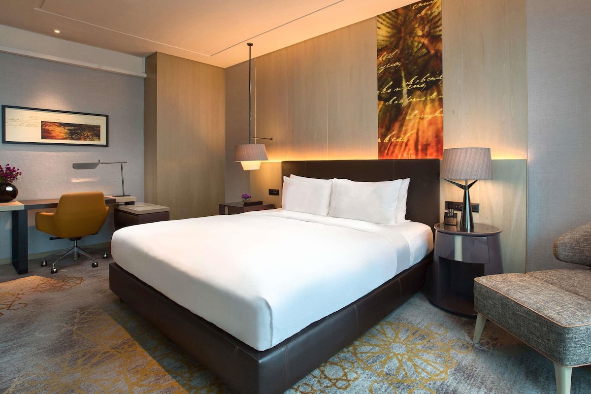 A modern hotel room with a large bed, bedside lamps, a desk with a chair, and a decorative wall piece.