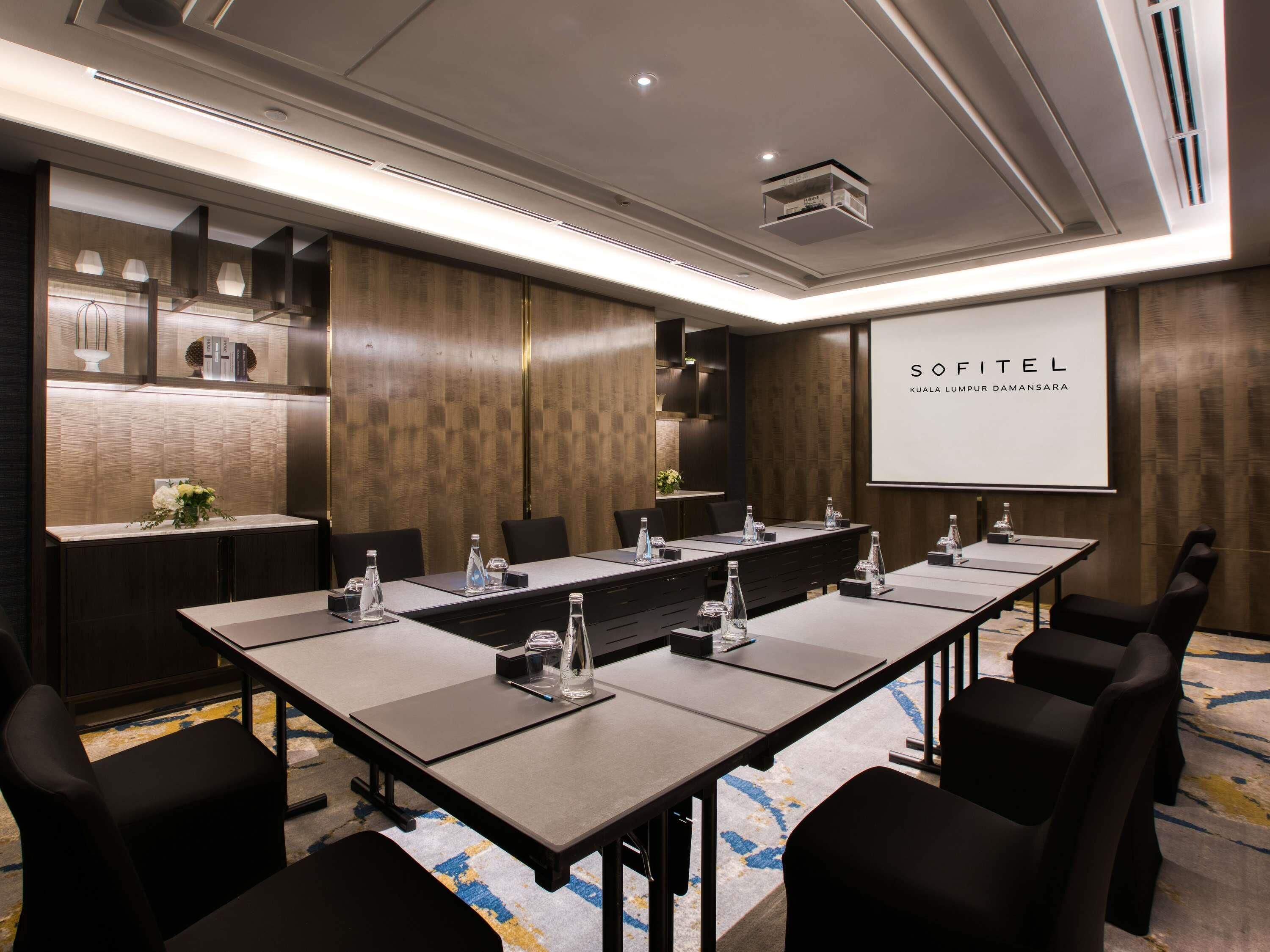 A conference room with a long table, chairs, a projector screen, and a wooden accent wall.