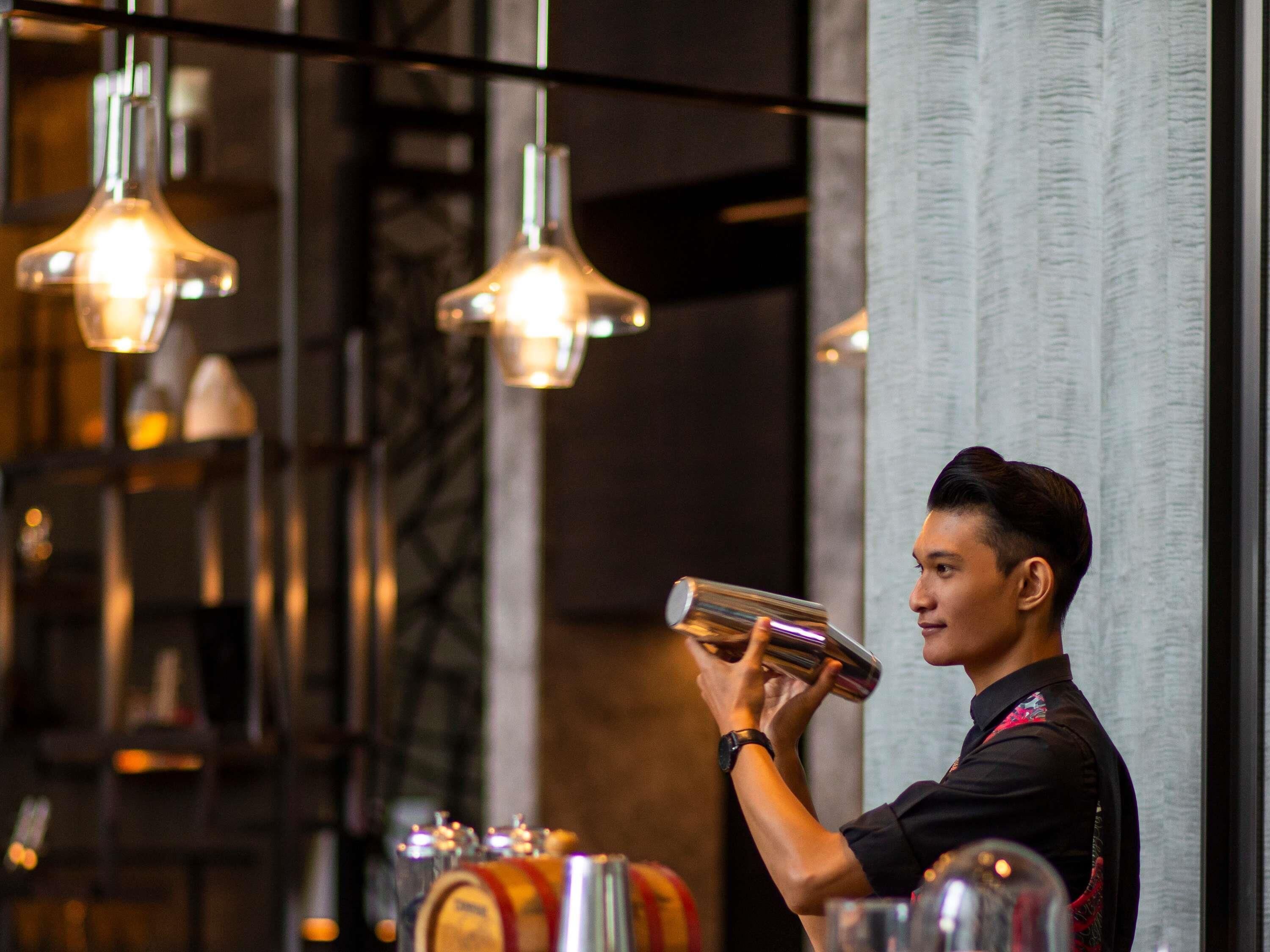 A bar area with hanging lights, a bartender preparing a drink, and a counter with various bottles and glasses.