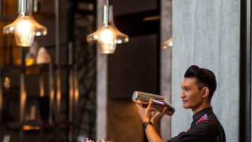 A bar area with hanging lights, a bartender preparing a drink, and a counter with various bottles and glasses.