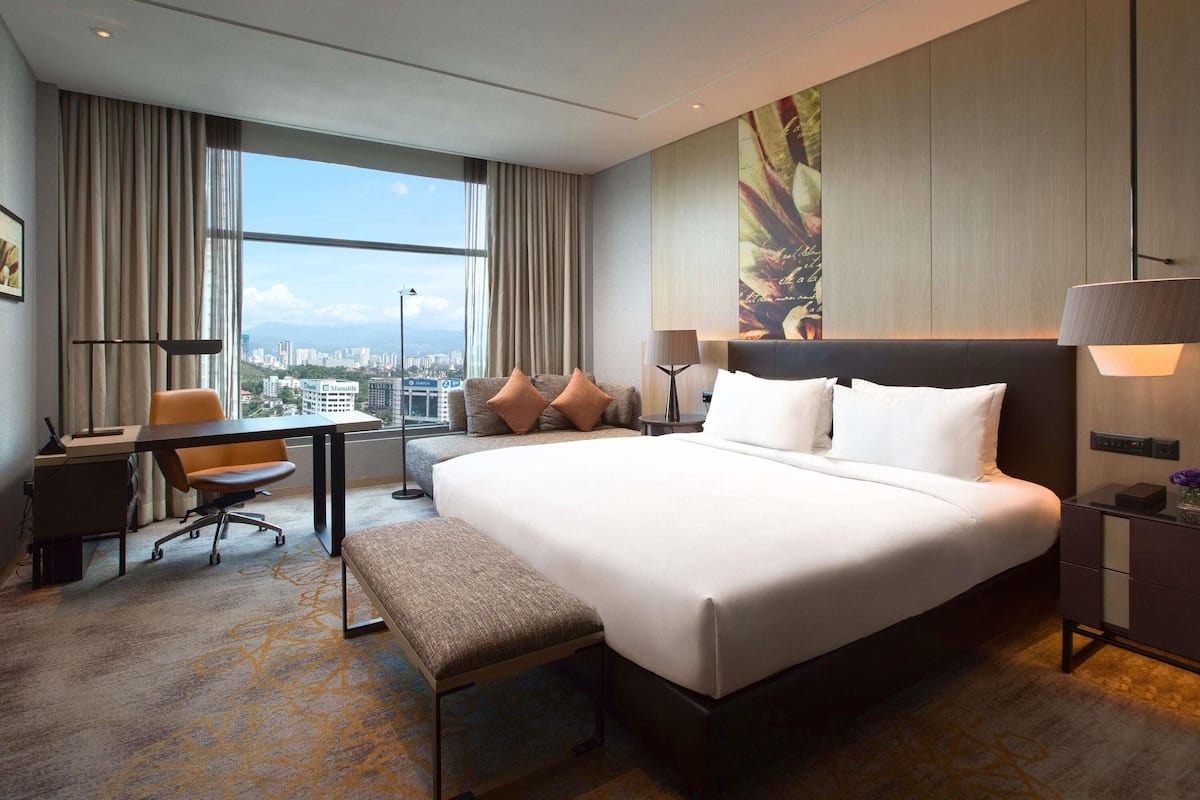 A hotel room with a large bed, a desk, a chair, a bench, a nightstand, and a view of the city through large windows.
