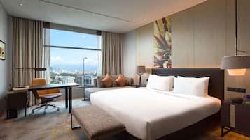 Habitación de hotel con una cama grande, un escritorio, una silla, un banco, una mesita de noche y vistas de la ciudad a través de amplios ventanales.