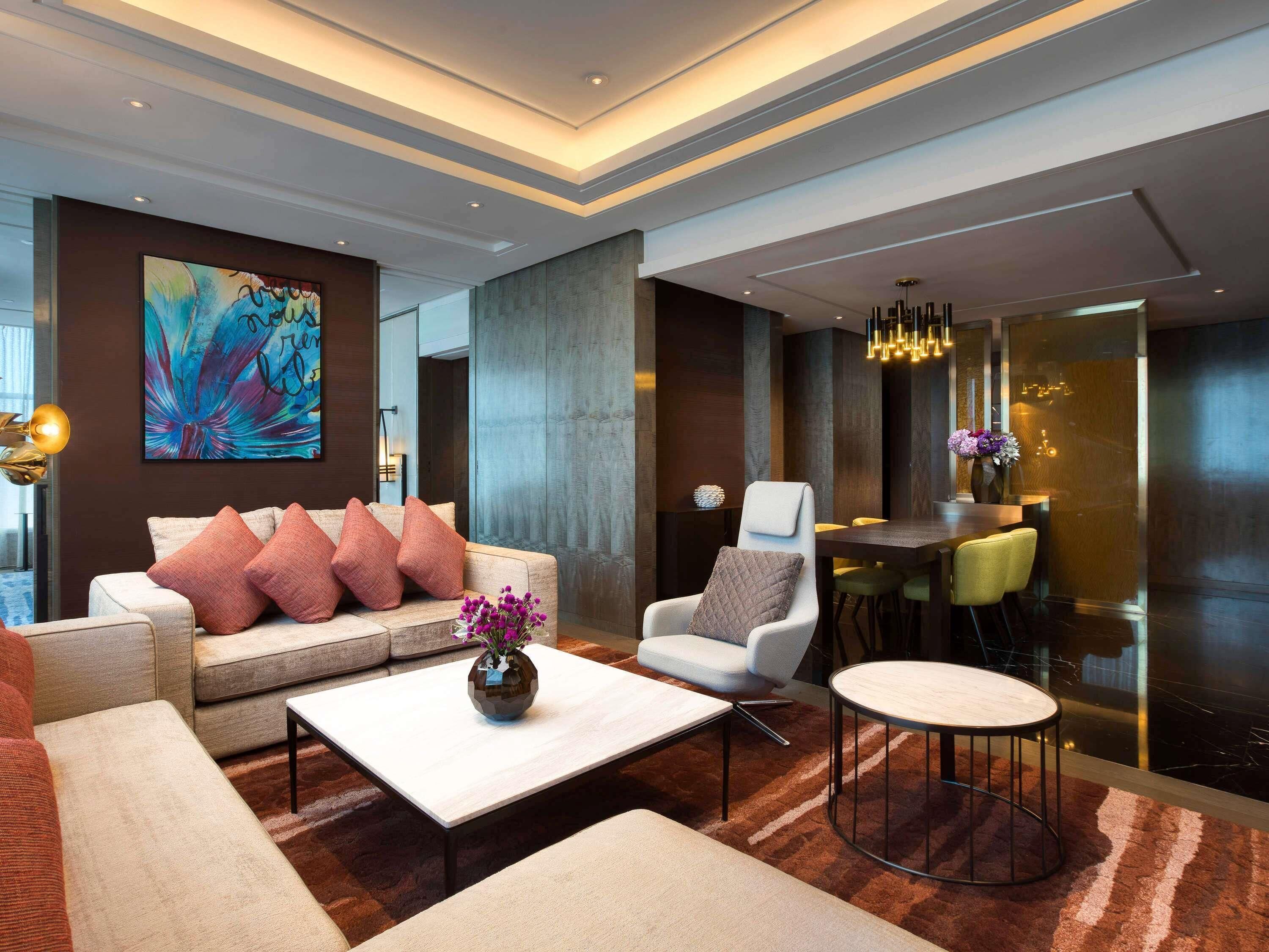 A modern hotel lobby with a sofa, armchairs, a coffee table, a dining area, and a large abstract painting on the wall.