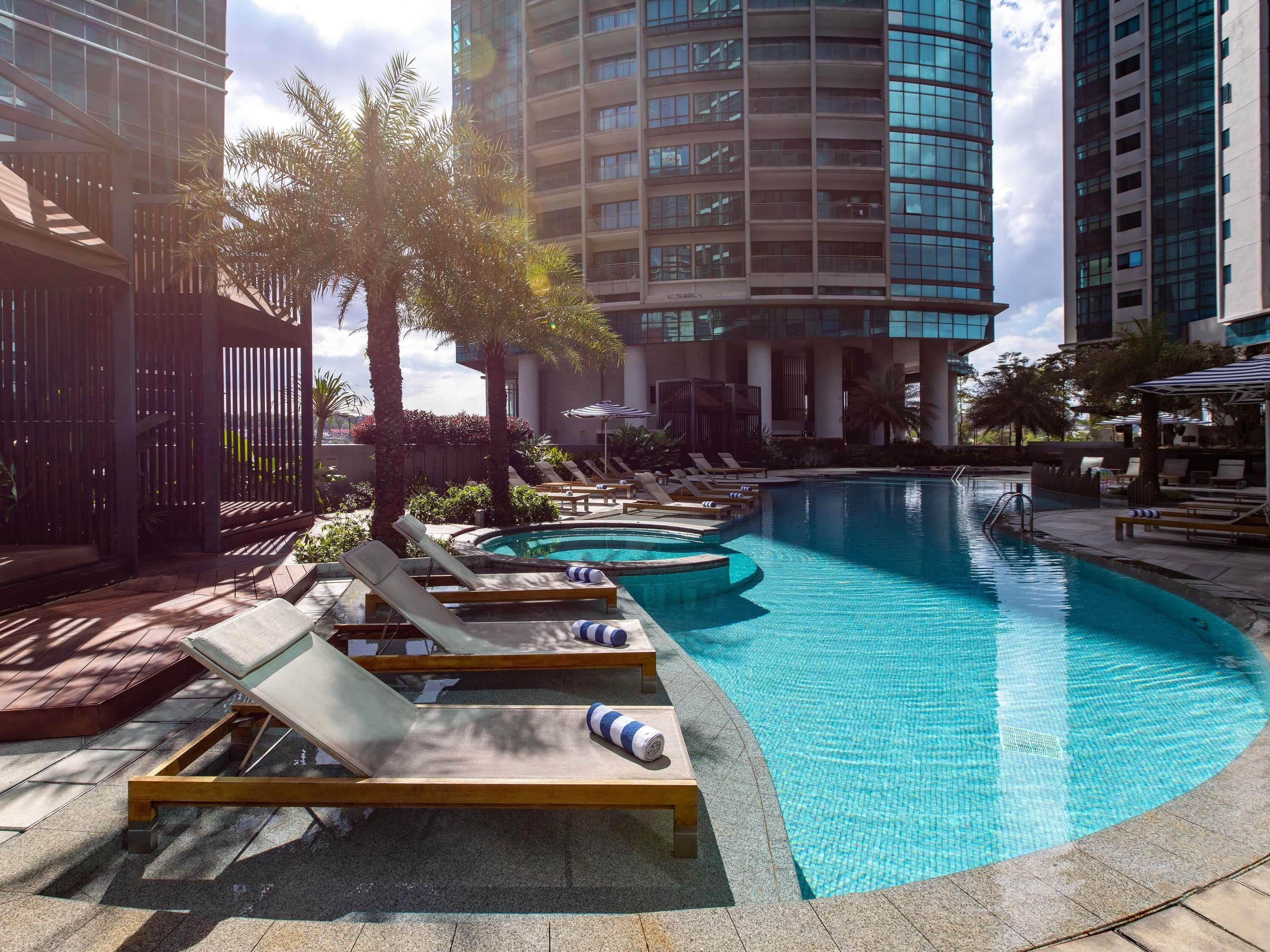 Une piscine entourée de chaises longues, de palmiers et, au loin, de bâtiments modernes.