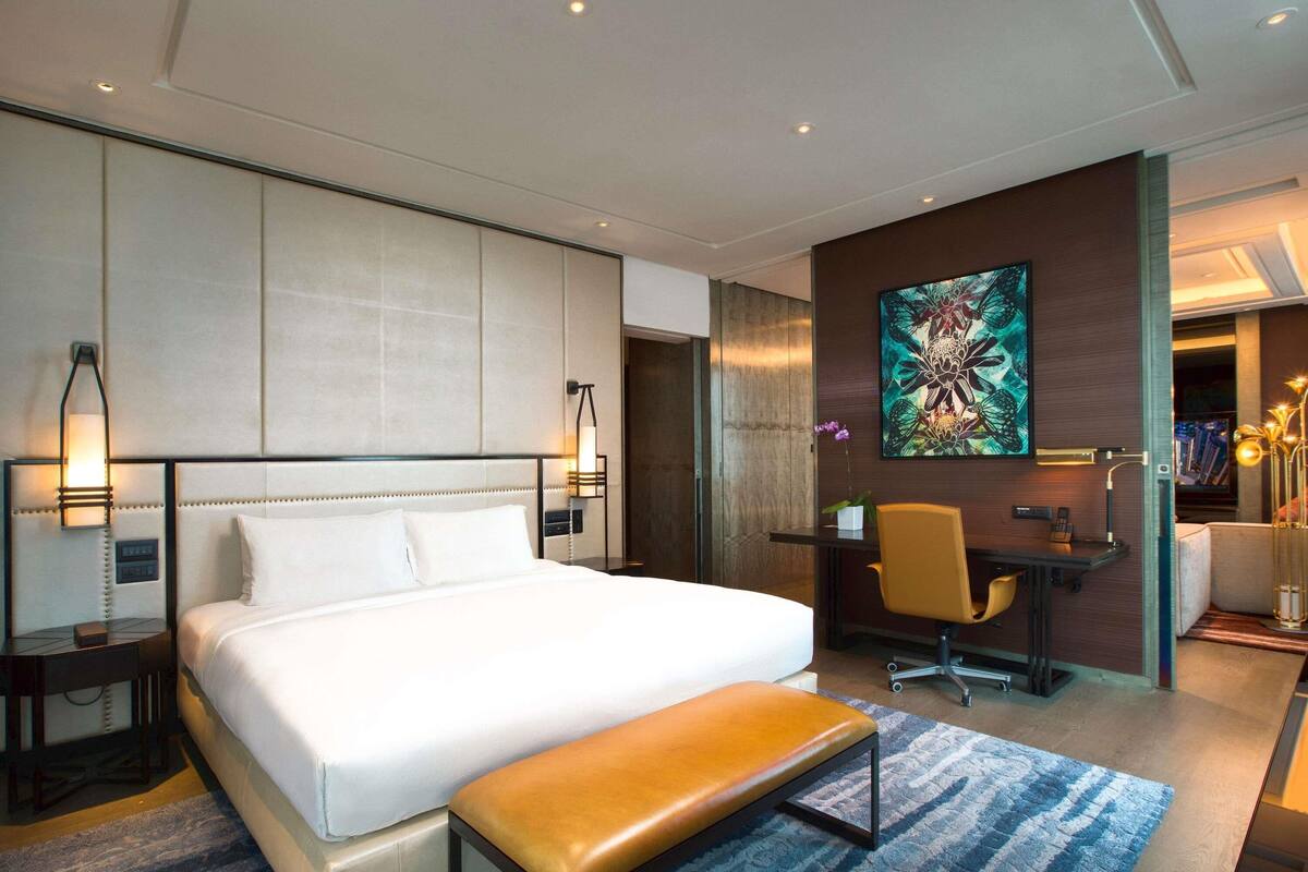A modern hotel room with a large bed, a desk, a chair, and a bench.