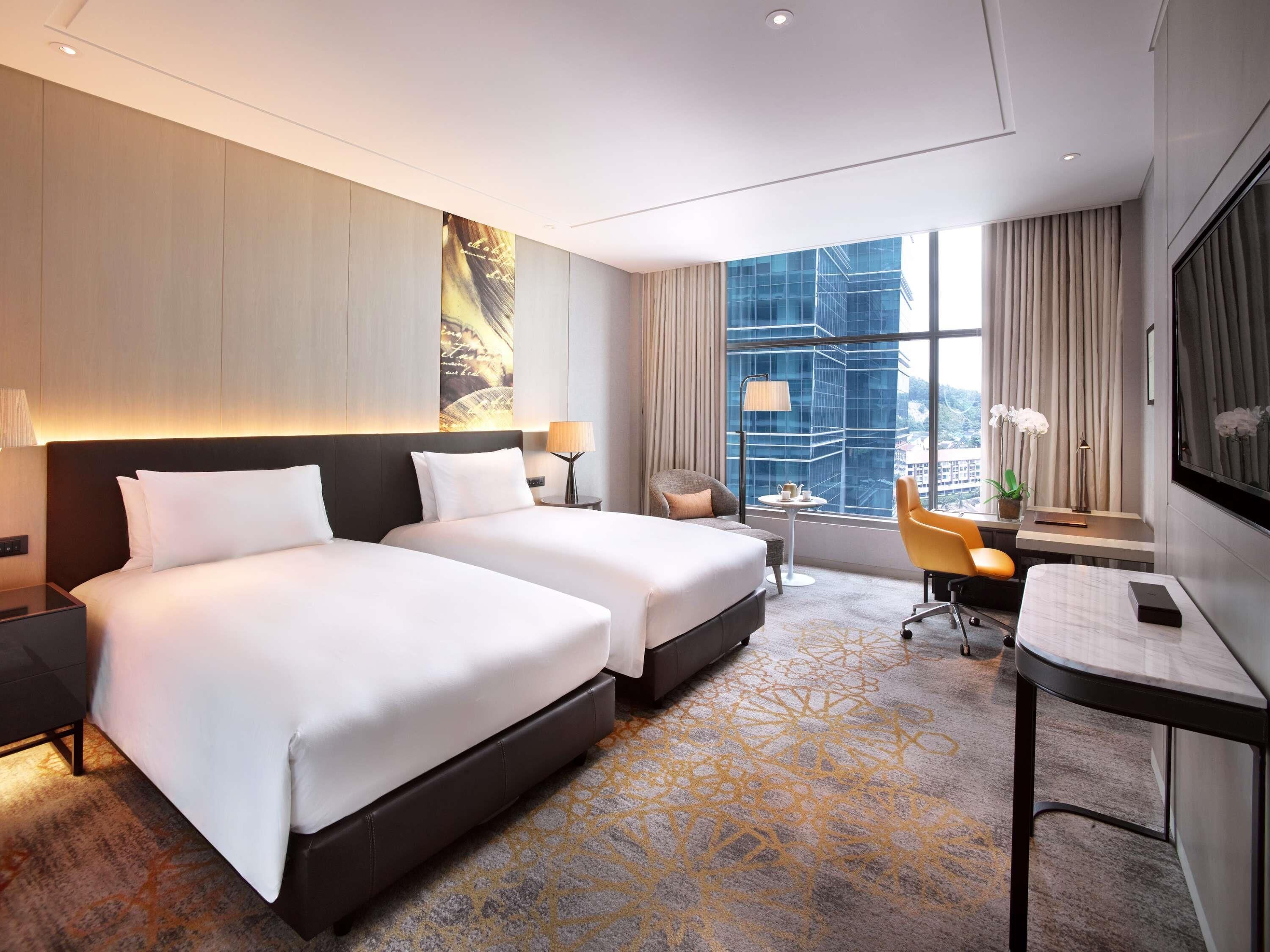 A hotel room with two beds, a desk, a chair, a lamp, and a large window with a city view.