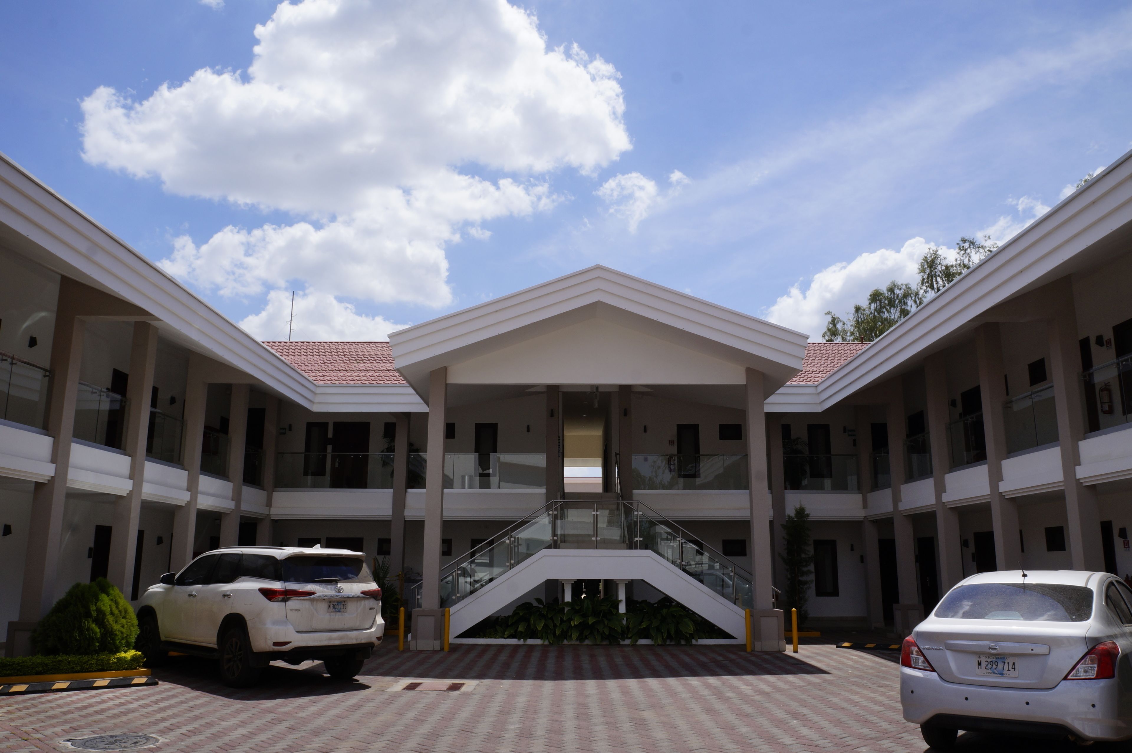 Un edificio de varios pisos con una escalera central, dos coches estacionados frente a él, y un cielo despejado con nubes.
